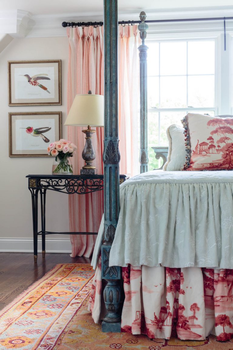 A cozy bedroom with a four-poster bed featuring green and pink bedding and a decorative skirt. A side table holds a cream lamp and a vase of pink roses. Two bird-themed framed artworks hang above. A window with pink curtains lets in natural light, and a colorful rug lies on the wooden floor.