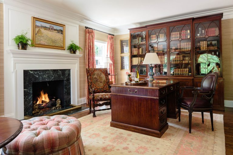 A cozy, elegant study features a dark wooden desk, an ornate armchair, and a round leather chair. A bookshelf with glass doors holds numerous books. A marble fireplace with a painting above it is lit, and two small plants adorn the mantel. The room is lit by natural light from a window with red curtains. A patterned rug covers the floor.