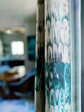Close-up of a curtain with a blue and white abstract pattern. The curtains texture is visible, featuring a blend of light and dark shades. The background is blurred, showing a softly lit room with a glimpse of furniture and a window.