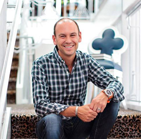 A smiling man with a shaved head sits on leopard-print stairs in a bright, modern space. He wears a black and white checkered shirt, jeans, and an orange watch. Behind him, theres a blurred staircase and a decorative chair with a club-shaped backrest.