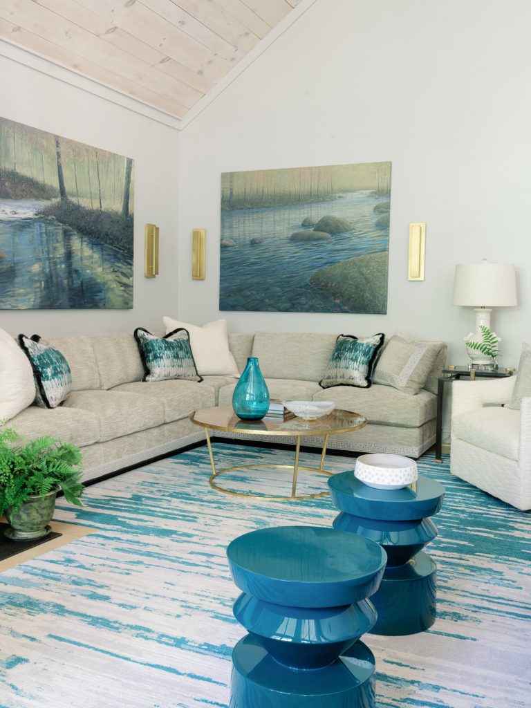 A modern living room features a beige sectional sofa with patterned blue and white cushions. Two large landscape paintings hang above. A round coffee table displays a blue vase. The room includes a teal patterned rug, two teal accent tables, a green plant, and a table lamp.