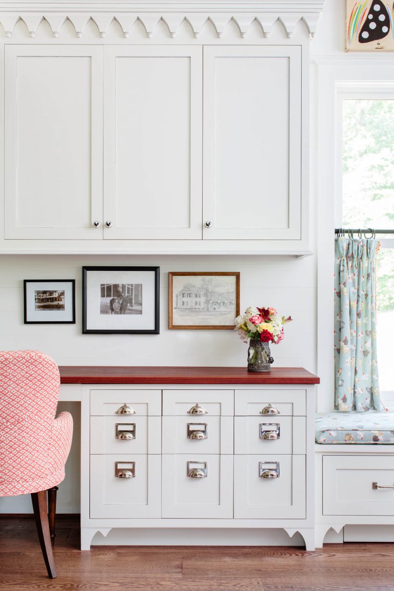 A cozy room with white cabinetry and a brown wooden countertop. Three framed pictures are displayed on the wall. A vase with colorful flowers sits on the countertop. A pink-patterned chair is partially visible. Nearby, a window with blue curtains and a cushioned bench lets in natural light.