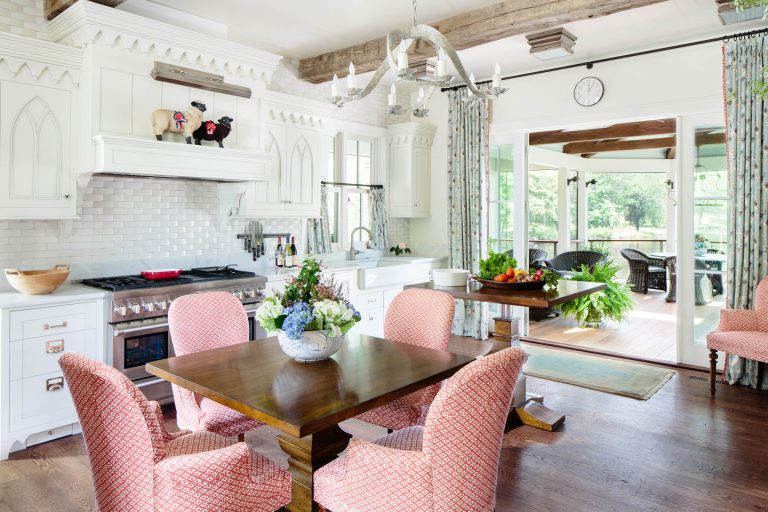 A bright kitchen with white cabinets and backsplash. A wooden dining table is surrounded by pink patterned chairs. A chandelier hangs above. There are flowers on the table, fruit on the counter, and a view of a sunroom with plants through a windowed door.