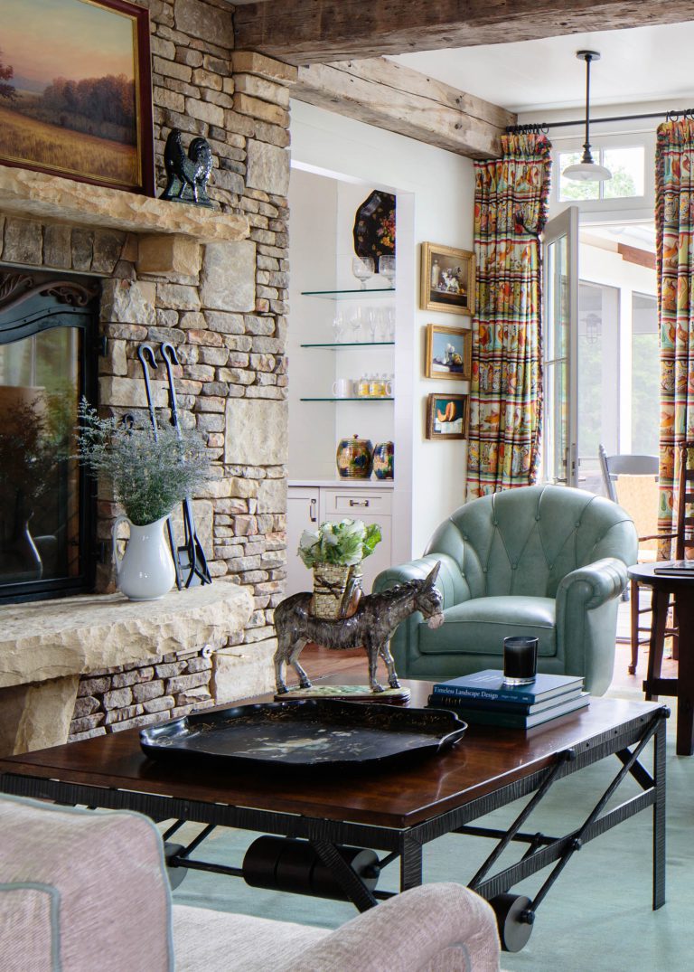 A cozy living room with a stone fireplace, a teal armchair, and colorful drapes. A wooden coffee table holds books, a black candle, and a decorative horse statue. Shelves with glassware are visible, and a door opens to an outdoor area. A rustic chandelier hangs above.