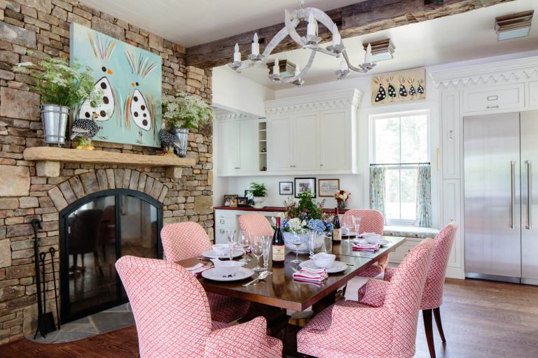 A cozy dining space with a wooden table set for six, featuring red patterned chairs. A stone fireplace with potted plants and art hangs on the left. A chandelier with a candle motif is overhead. The kitchen area in white features cabinets and a large fridge, with a window view to the right.