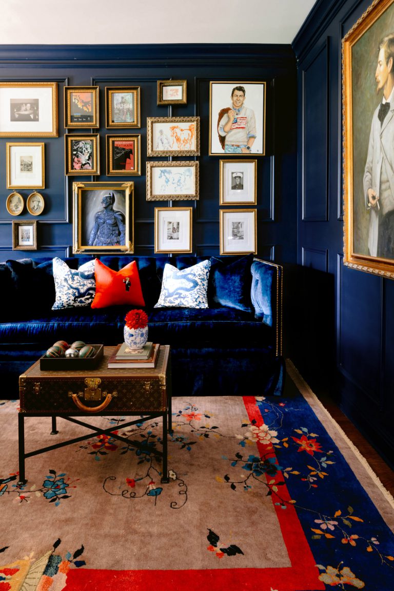 A sophisticated living room features deep blue walls adorned with framed art in gold frames. A plush navy sofa with patterned cushions is placed against the wall. A vintage trunk serves as a coffee table on a floral-patterned rug, adding an elegant touch to the space.