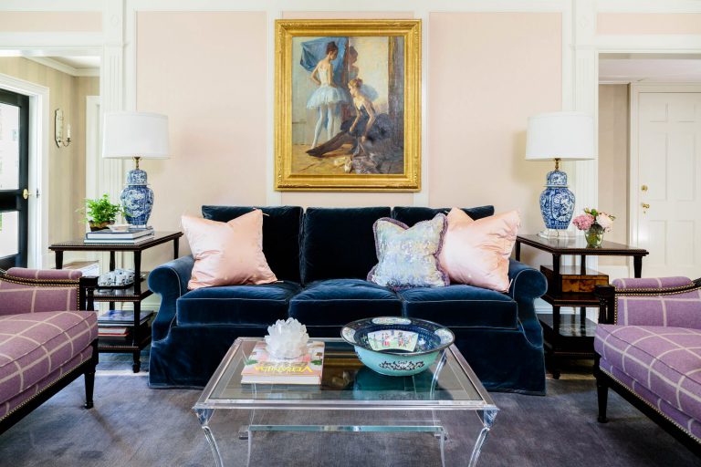 A living room with a dark blue velvet sofa adorned with pink and floral pillows. Above it, a framed painting of ballet dancers hangs on a pale pink wall. Flanking the sofa are two tables with blue and white lamps. A glass coffee table holds books, a crystal, and decorative bowls.