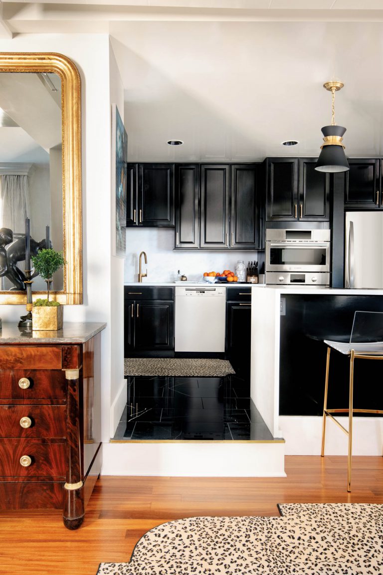 Elegant kitchen scene with black cabinets and stainless steel appliances. A modern pendant light hangs above a gold-accented bar stool. A decorative rug partially covers the wooden floor, and a large mirror with a gold frame is positioned next to a wooden dresser with ornate details.