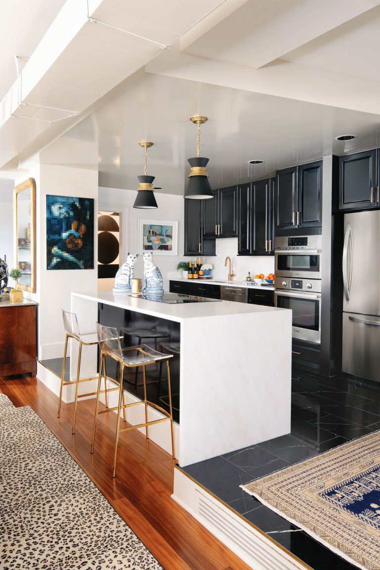 Modern kitchen with black cabinetry and stainless steel appliances. A white marble island features two gold-framed clear chairs. Above the island hang two black and gold pendant lights. The floor is a mix of dark tiles and wood with a leopard print rug. Art and decor add color to the space.