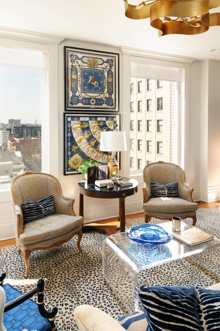 A bright living room with two ornate chairs and a round wooden table between them, topped with a lamp and books. Behind are two patterned wall hangings. The room includes a clear coffee table, a blue vase, animal print rug, and large windows overlooking city buildings.