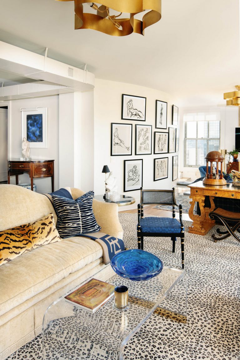 Stylish living room with a beige sofa adorned with zebra and tiger print cushions. A clear acrylic coffee table displays a blue glass bowl and a book. Gold ceiling fixtures add elegance. The room features a wall of abstract framed art, leopard print carpet, and wooden furniture.