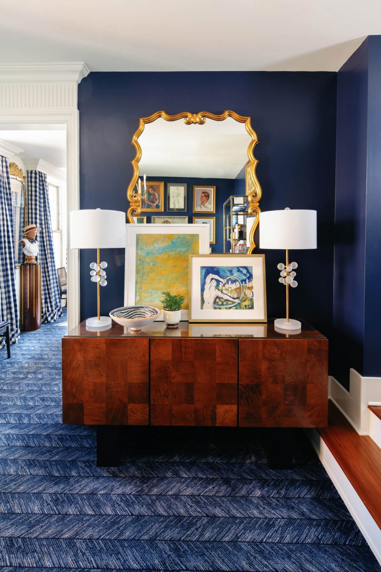A stylish room features a dark wooden sideboard with two white lamps, abstract art pieces, and a potted plant. A gold-framed mirror above reflects more artwork. The navy blue walls and patterned carpet complement the decor, enhancing the rooms elegant and contemporary feel.