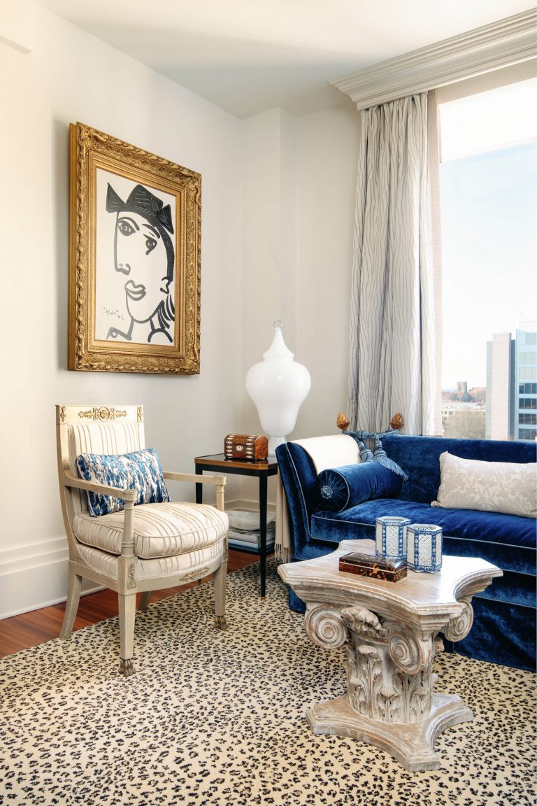 A sophisticated living room with a blue velvet sofa, embellished with pillows, sits on a leopard print rug. A baroque-style chair and ornate side table with decorative boxes are present. A large abstract painting in a gold frame hangs above. A white vase is near the window with beige curtains.