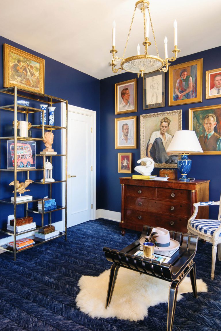 A stylish room with deep blue walls adorned with various framed portraits. A brass and glass shelving unit holds books and decor, including a bust. A wooden dresser topped with a lamp and sculpture stands against the wall. A small table with a hat and books sits on a white rug over a blue carpet.