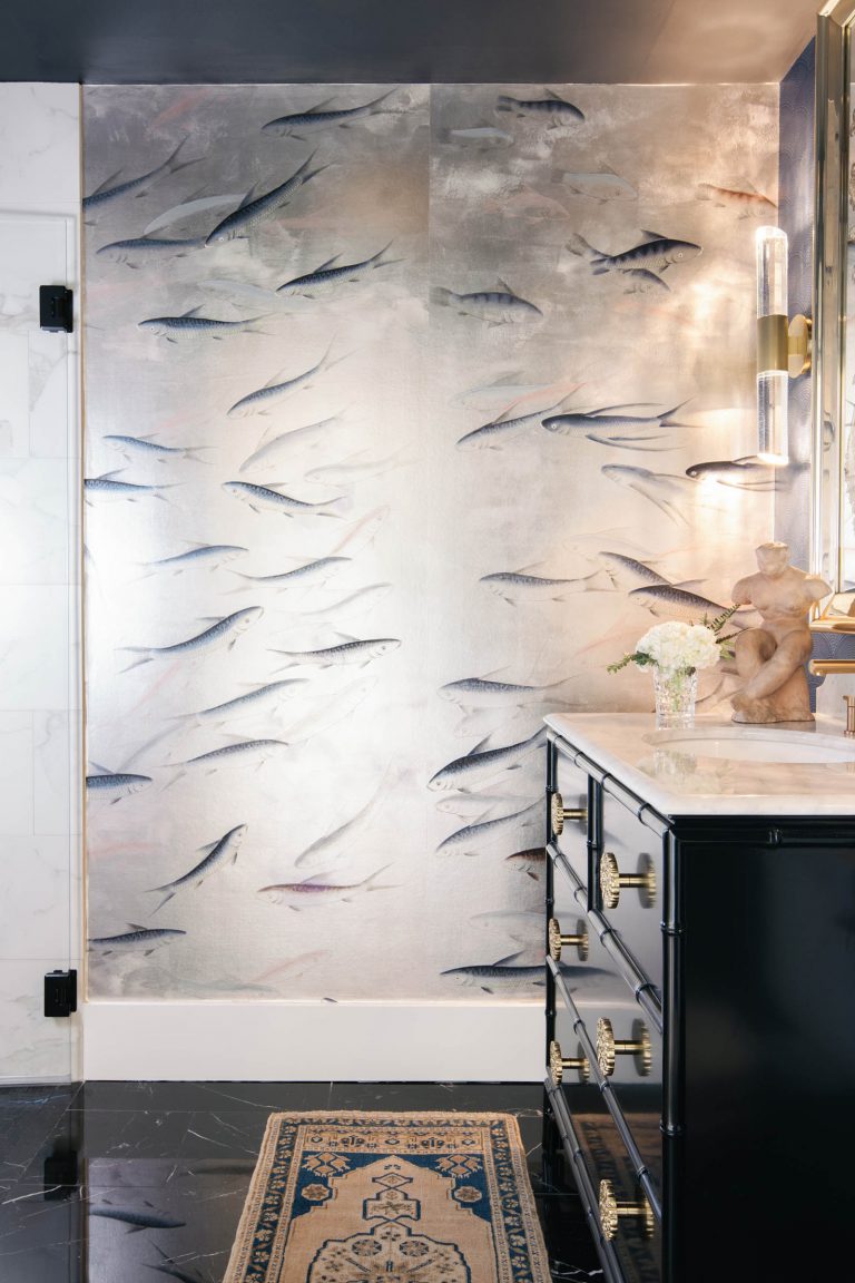 Bathroom with a metallic fish-motif wallpaper, featuring flying fish. A dark vanity with gold accents sits underneath a marble countertop. On the counter, there’s a small vase with flowers and a decorative sculpture. The floor is black marble with a patterned rug.