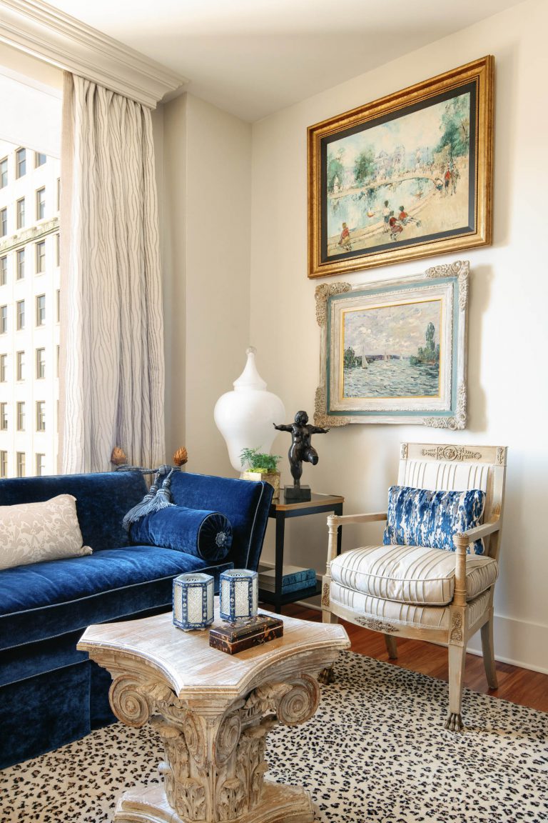 Elegant living room with a deep blue velvet sofa and a decorative beige cushion. A vintage armchair with a blue patterned cushion sits nearby. Two framed paintings adorn the walls above. A unique carved wooden coffee table holds two patterned lanterns. A large white vase stands in the corner.