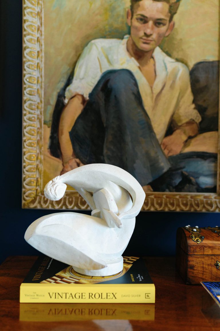A white abstract sculpture rests on a book titled Vintage Rolex. In the background, theres a painting of a person with brown hair, wearing a white shirt and blue pants, sitting against a dark background. A wooden box with metal details sits beside the sculpture on the wooden surface.