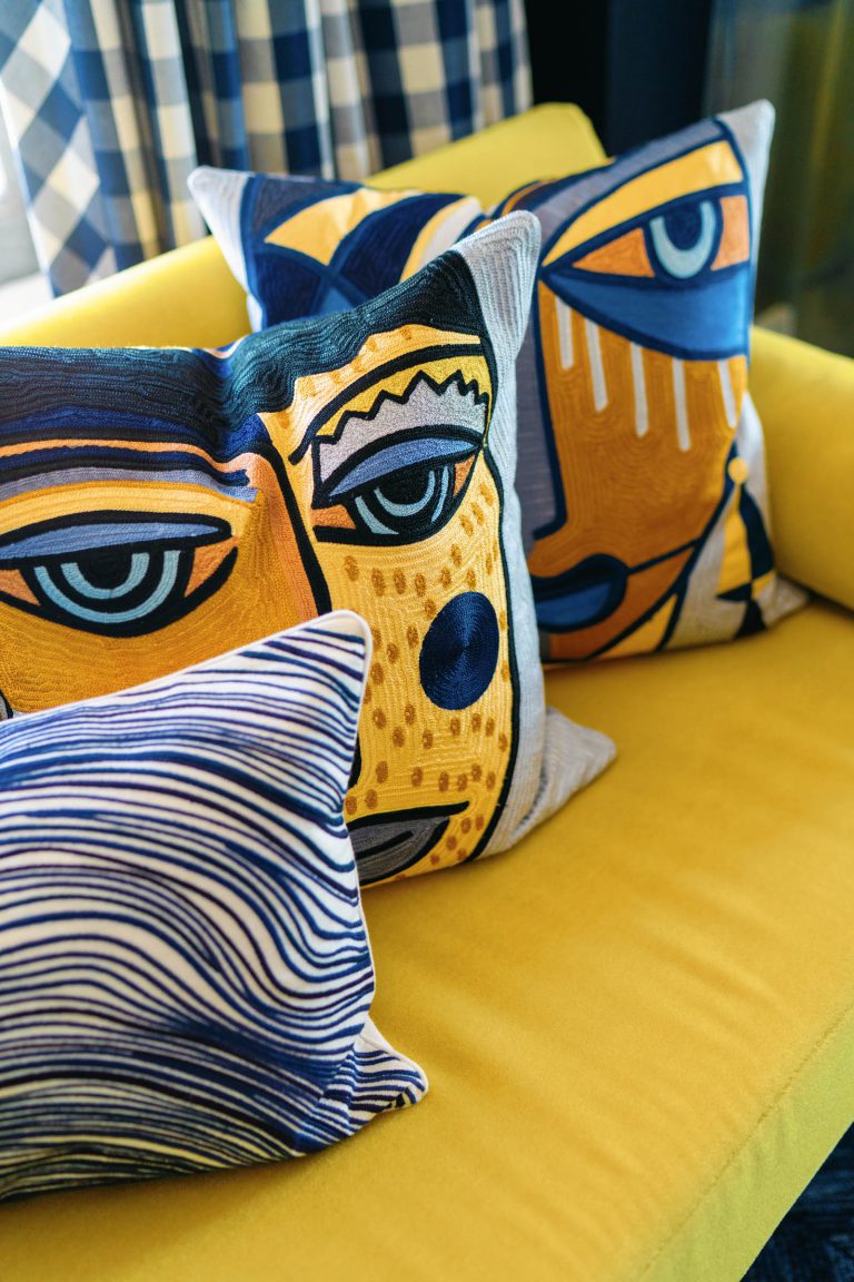 Three decorative pillows with abstract face designs rest on a bright yellow sofa. The front pillow features wavy blue lines, while the two behind it display stylized faces in blue and yellow tones. A checkered curtain is visible in the background.