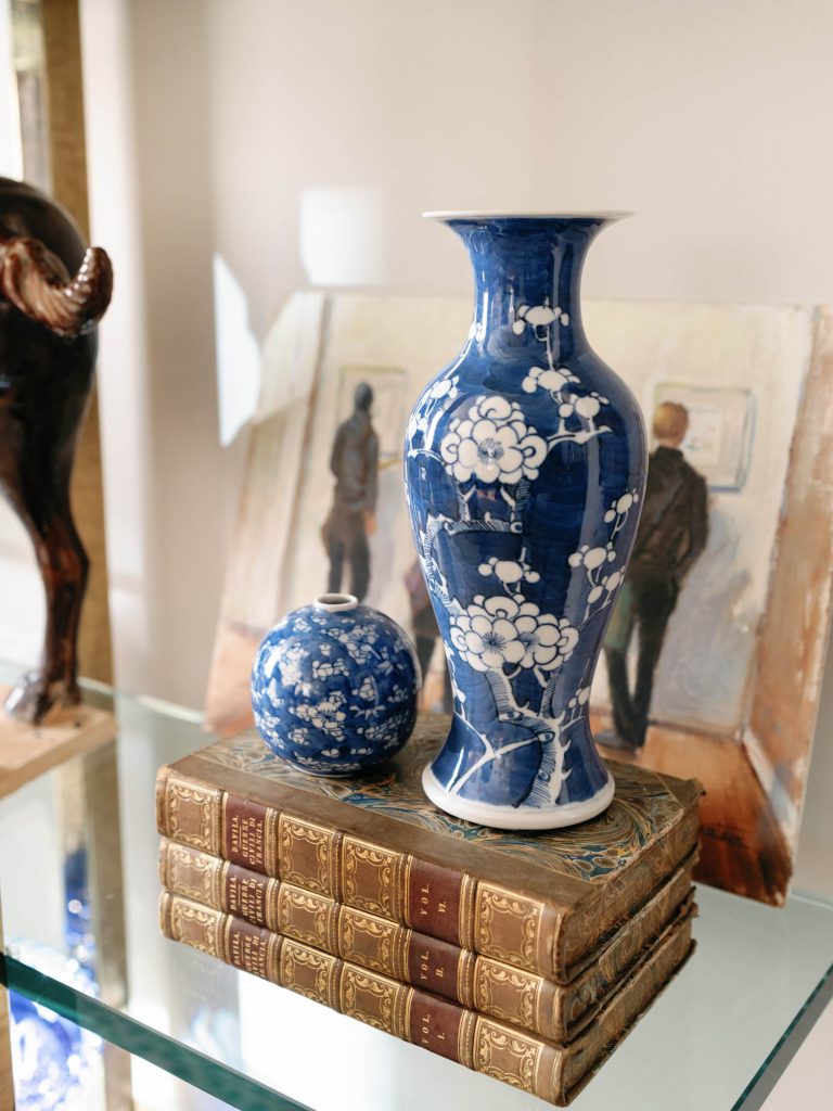 A blue and white floral vase and a round porcelain object sit on a stack of old, leather-bound books. Behind them, a painting of two figures in a gallery is displayed on a glass shelf. An animal sculpture is partially visible on the left. The setting is elegant and artistic.