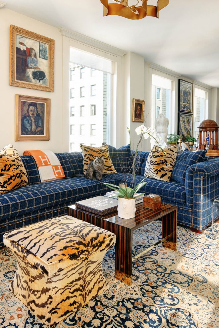 A stylish living room features a blue plaid sectional sofa adorned with orange and patterned pillows. A marble coffee table sits atop a patterned rug. Artworks decorate the walls. Large windows let in natural light. A unique stool with a tiger print fabric adds flair to the space.