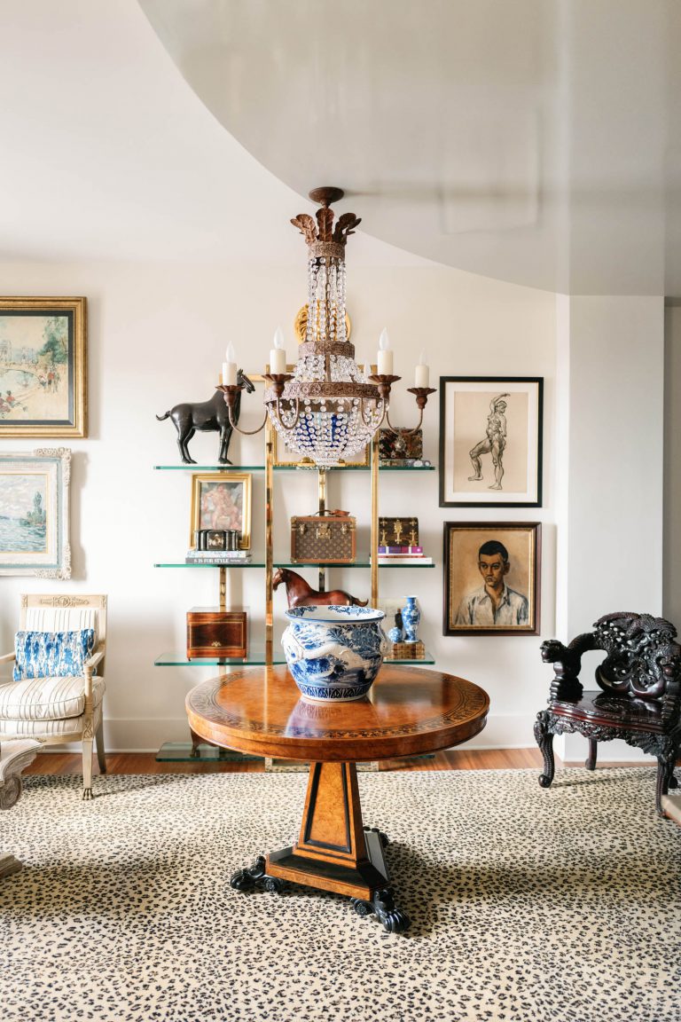 Elegant living room with a round wooden table displaying a blue and white porcelain vase. An ornate chandelier hangs above. The wall features framed art and shelves with decorative pieces, including an elephant figurine. Richly textured armchairs and a leopard-patterned carpet complete the scene.