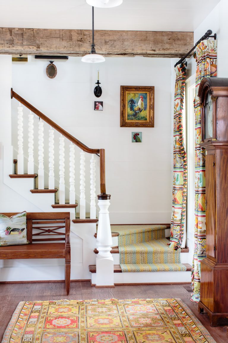 A cozy interior with a wooden staircase featuring white spindles and a brown railing. An antique grandfather clock stands to the right. Colorful patterned curtains and a matching rug add vibrancy. Artwork hangs on the walls, and a wooden bench with cushions is on the left.