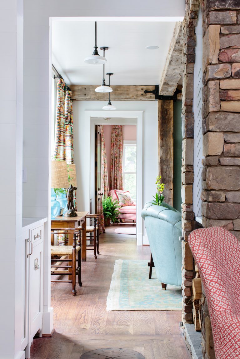 A cozy, well-lit hallway features rustic stone walls and wooden beams. The area includes a blue armchair, patterned curtains, a small sitting area with a lamp, and a red cushioned chair. Wooden flooring and a small green rug add warmth, with vibrant plants visible near the windows.