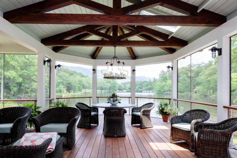 A spacious gazebo with wooden floors and a vaulted ceiling features wicker furniture around a central round table. A chandelier hangs above, and large screened windows overlook a serene lakeside view with trees and mountains in the background. Potted plants add a touch of greenery.