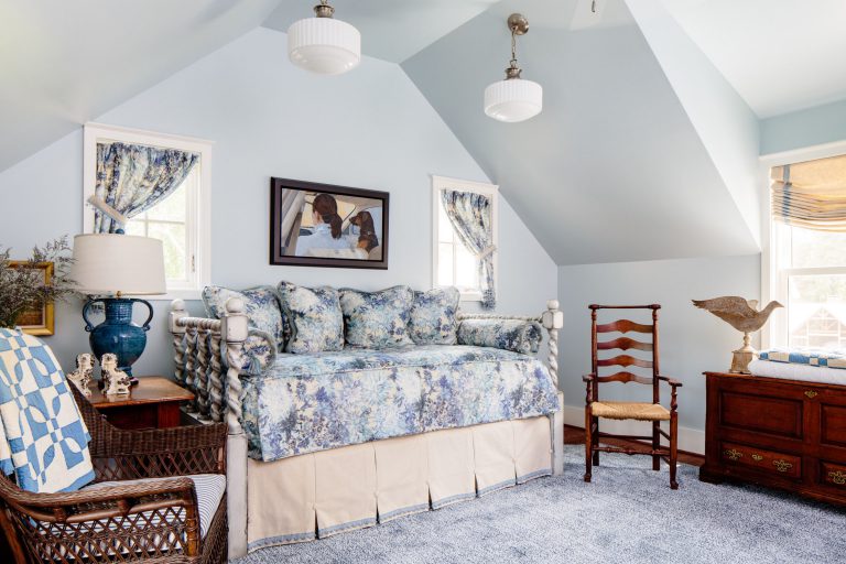 A cozy room with light blue walls features a daybed adorned with floral cushions and a beige skirt. Above it, a framed photo decorates the space. Two small windows have floral curtains. A wooden chair sits beside a chest topped with a wooden sculpture. Two ceiling lights illuminate the room.