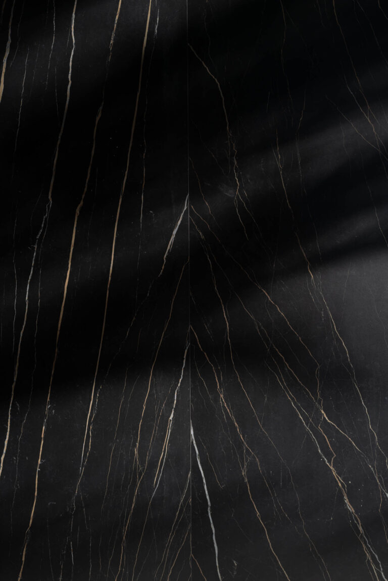 A textured black marble surface with subtle brown and white veins running diagonally across it. Soft shadows create a gradient from top right to bottom left, adding depth and contrast to the stones smooth, polished appearance.