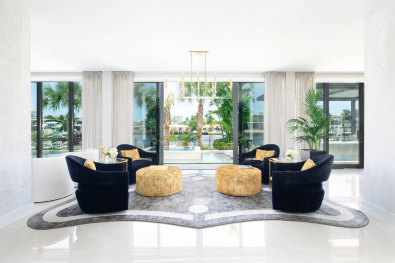 A modern, bright lounge area with a white floor and walls features four black, curved armchairs and two round yellow ottomans on a gray abstract rug. Large windows with sheer curtains overlook a tropical outdoor scene with palm trees and water. A sleek, gold ceiling light is centered above.