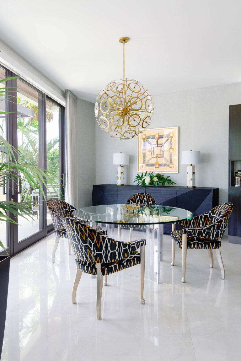 A modern dining room with a round glass table and four patterned chairs. Above the table hangs an intricate gold chandelier. A sideboard with two lamps and a framed abstract art piece sits against the wall. Large windows overlook lush greenery, and the floor is glossy white tile.