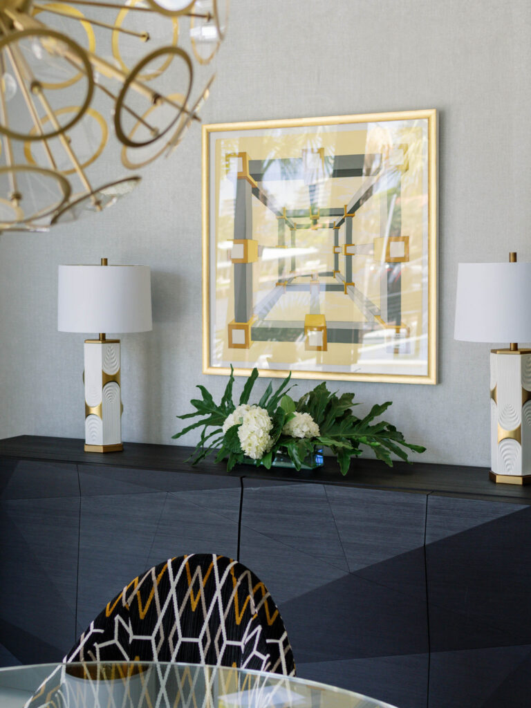 A modern dining room features a sleek black sideboard with two geometric-patterned lamps. A framed abstract artwork hangs above. A floral arrangement of white hydrangeas sits in the center. A round glass table with patterned chairs is in the foreground, beneath a gold chandelier.