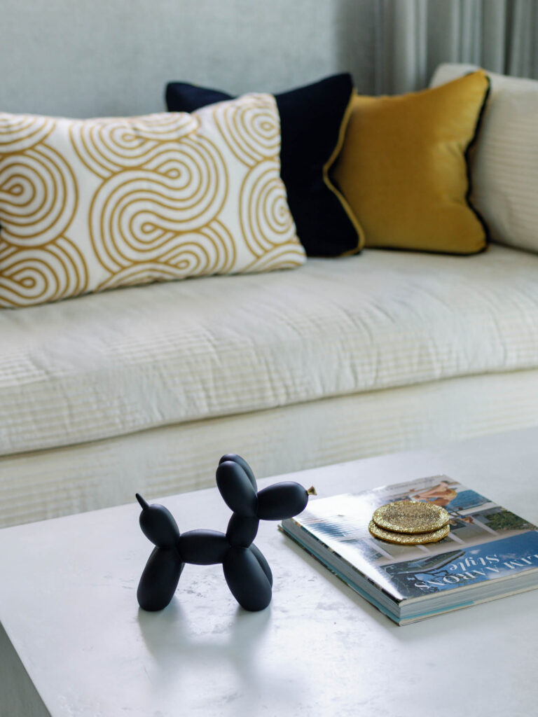 A light-colored sofa with patterned and solid pillows is in the background. In the foreground, a black balloon dog sculpture sits on a white table next to stacked magazines and a small, round gold purse. The scene is elegantly arranged in a modern setting.
