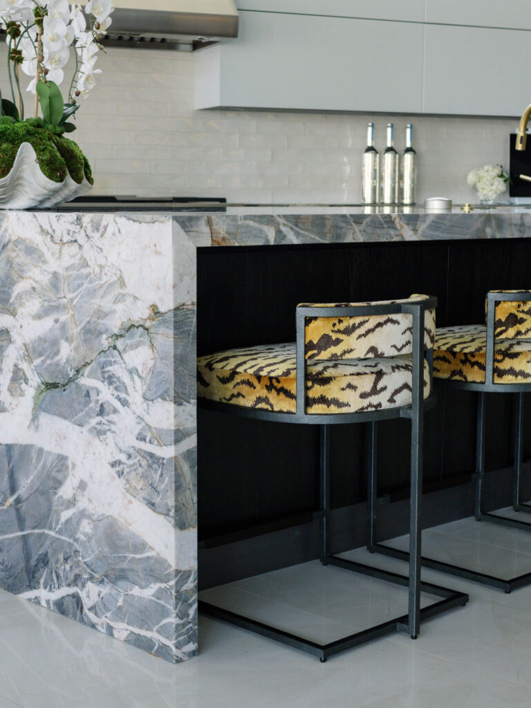 A modern kitchen features a marble island with gray, white, and green veining. Two barstools with black metal frames and tiger-patterned cushions are tucked under the counter. A large decorative shell with orchids sits on the island, and three bottles are lined up in the background.