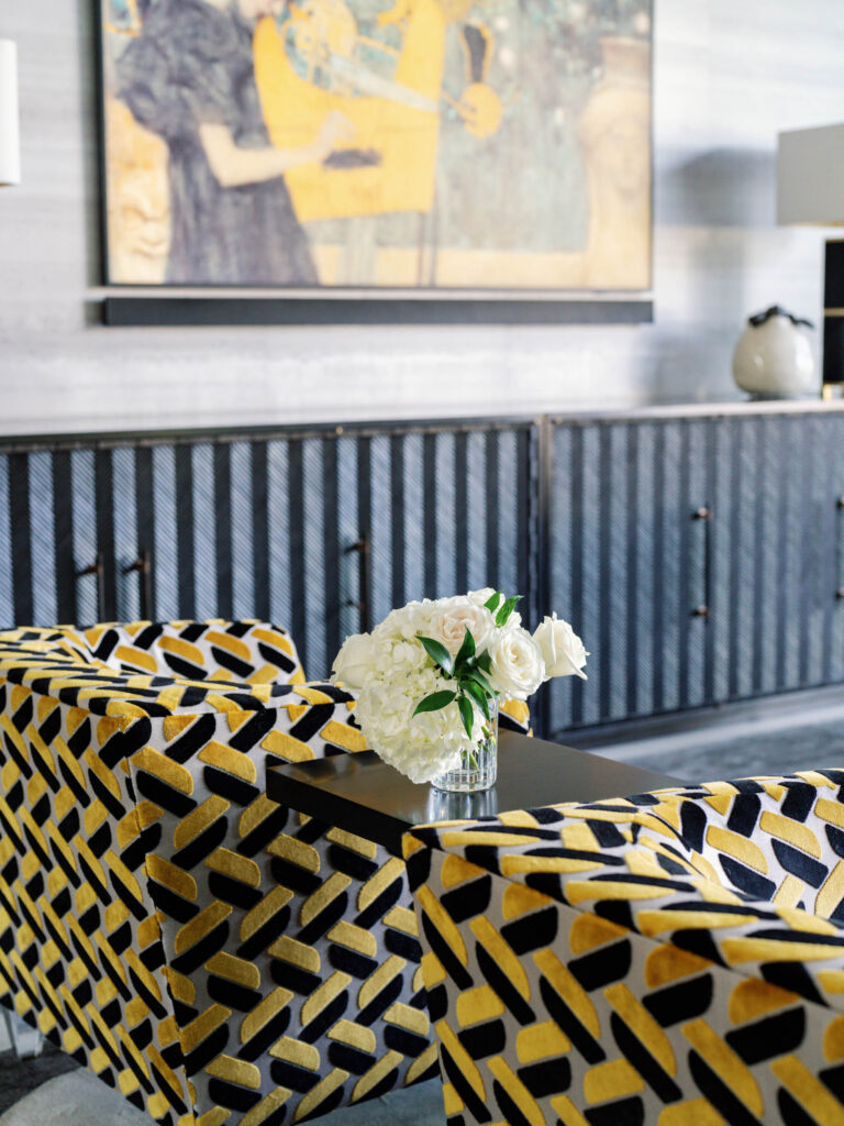 A modern living room features two yellow and black patterned armchairs. A glass vase with white flowers sits on a table between them. In the background, there are dark cabinets and a large framed painting with abstract figures on the wall. A soft gray carpet completes the scene.