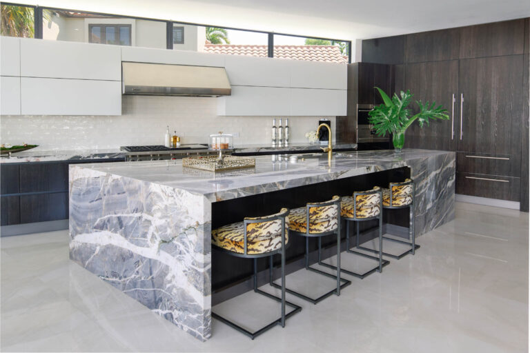 Modern kitchen with a large marble island featuring gray and white swirls. Four stools with patterned upholstery are at the island. Dark wood and white cabinetry contrast with stainless steel appliances. A gold faucet and green plant adorn the counter. Natural light fills the space.