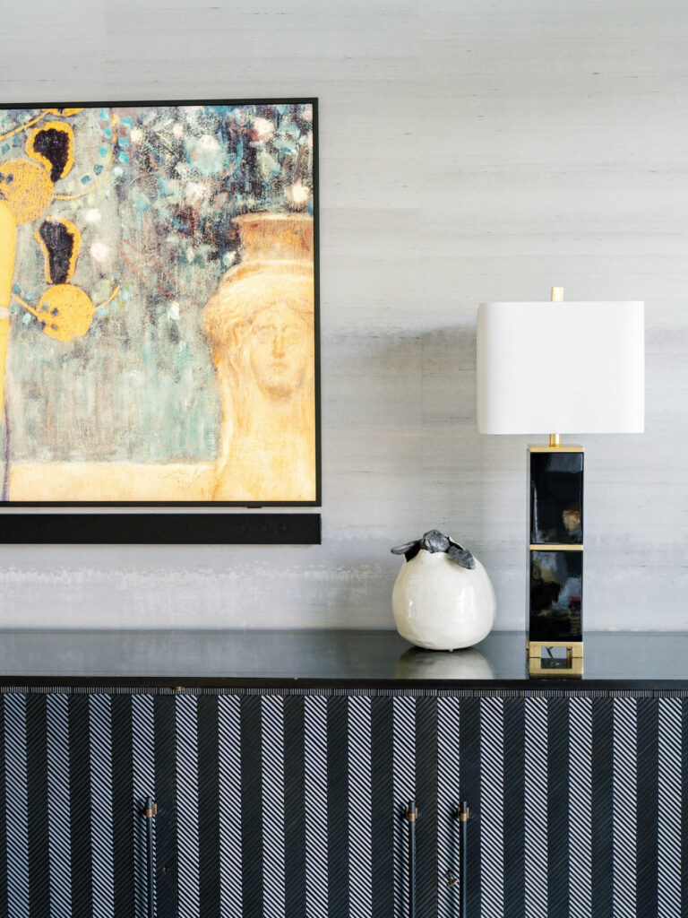 A modern interior features a striped black and white sideboard with a white vase and a black and gold table lamp. Above, a framed abstract artwork with muted colors is mounted on a textured gray wall, depicting a partial portrait and abstract shapes.