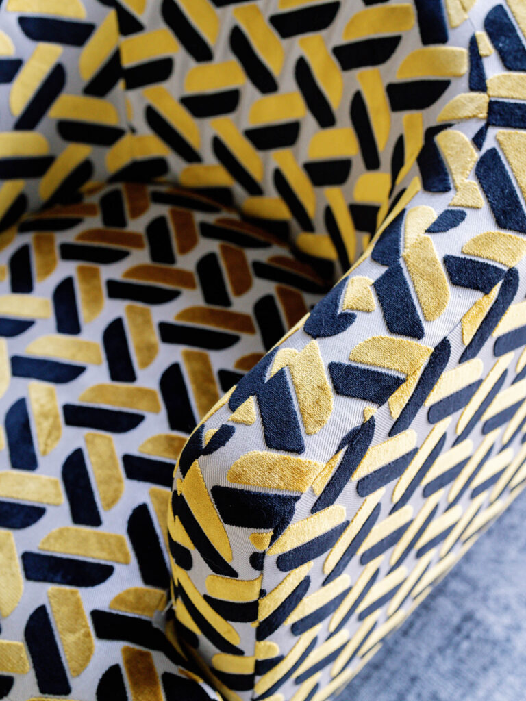 A close-up of a fabric chair with a bold geometric pattern. The upholstery features interlocking shapes in yellow, gold, and black on a white background. The chair sits on a gray carpet, and the angle focuses on the armrest and seat cushion.