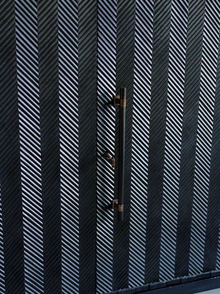 Close-up of a textured dark metal door with diagonal herringbone patterns. A sleek, vertical handle with a metallic finish is positioned centrally, adding a modern touch to the design. The patterned surface reflects light subtly, enhancing the depth of the texture.