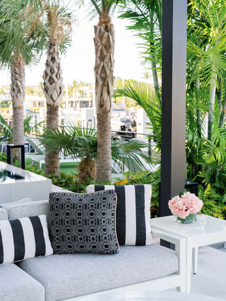 A stylish outdoor patio features a grey sofa adorned with black and white patterned cushions. A small white table beside the sofa holds a bouquet of pink flowers. Lush green palm trees and plants surround the area, creating a tropical ambiance. A view of parked vehicles is visible in the background.