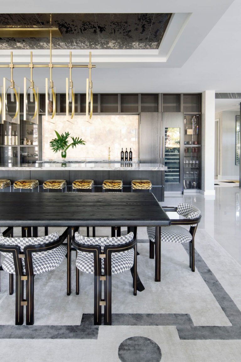Modern dining room with a large black table and eight patterned chairs. A stylish gold chandelier hangs above. In the background, a sleek kitchen features dark cabinetry, a stone backsplash, and a vase with green leaves. A wine fridge and a polished floor enhance the contemporary look.