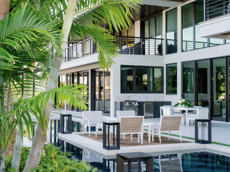 Modern outdoor patio with white furniture, including tables and chairs, set around a sleek rectangular pool. The building features large glass windows and balconies with black railings. Tropical plants with green fronds frame the foreground, adding a lush touch to the scene.