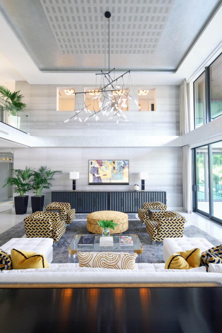 A modern, spacious living room features a high ceiling with a geometric chandelier. The room has patterned mustard and black chairs around a glass coffee table, with a large beige ottoman. A painting and potted plants decorate the back wall. The room has ample natural light and elegant decor.