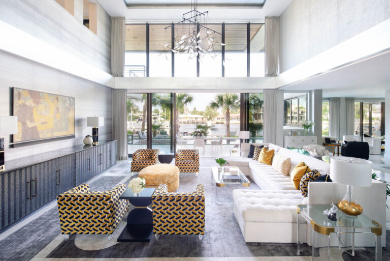Spacious modern living room with high ceilings, large windows, and abundant natural light. Features a white sectional sofa with yellow cushions, four patterned armchairs, and a round yellow ottoman. A chandelier hangs above; outside, palm trees are visible through glass doors.
