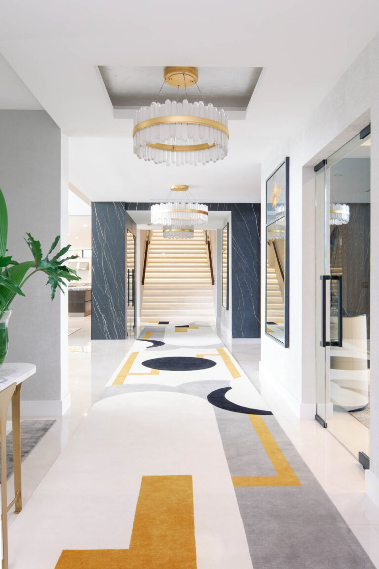 A modern hallway features a colorful geometric carpet with white, black, gray, and mustard patterns. Elegant chandeliers hang from the ceiling. A large plant is on a table to the left. Marble stairs with wooden railings ascend in the background. Glass doors are on the right.