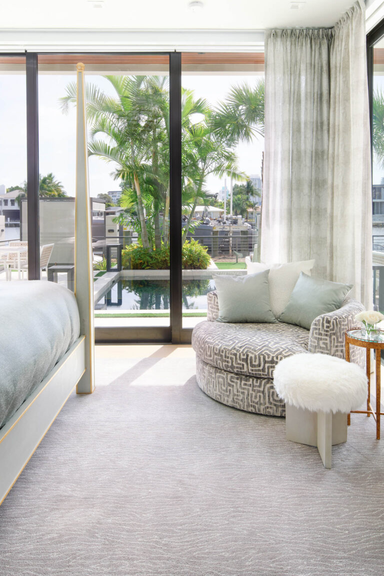 Bright bedroom with large windows overlooking a lush outdoor view. A cozy corner features a patterned circular chair, light blue and gray pillows, and a small stool with a fluffy white top. The bed has a light blue cover, and sheer curtains add an airy feel. Palm trees are visible outside.