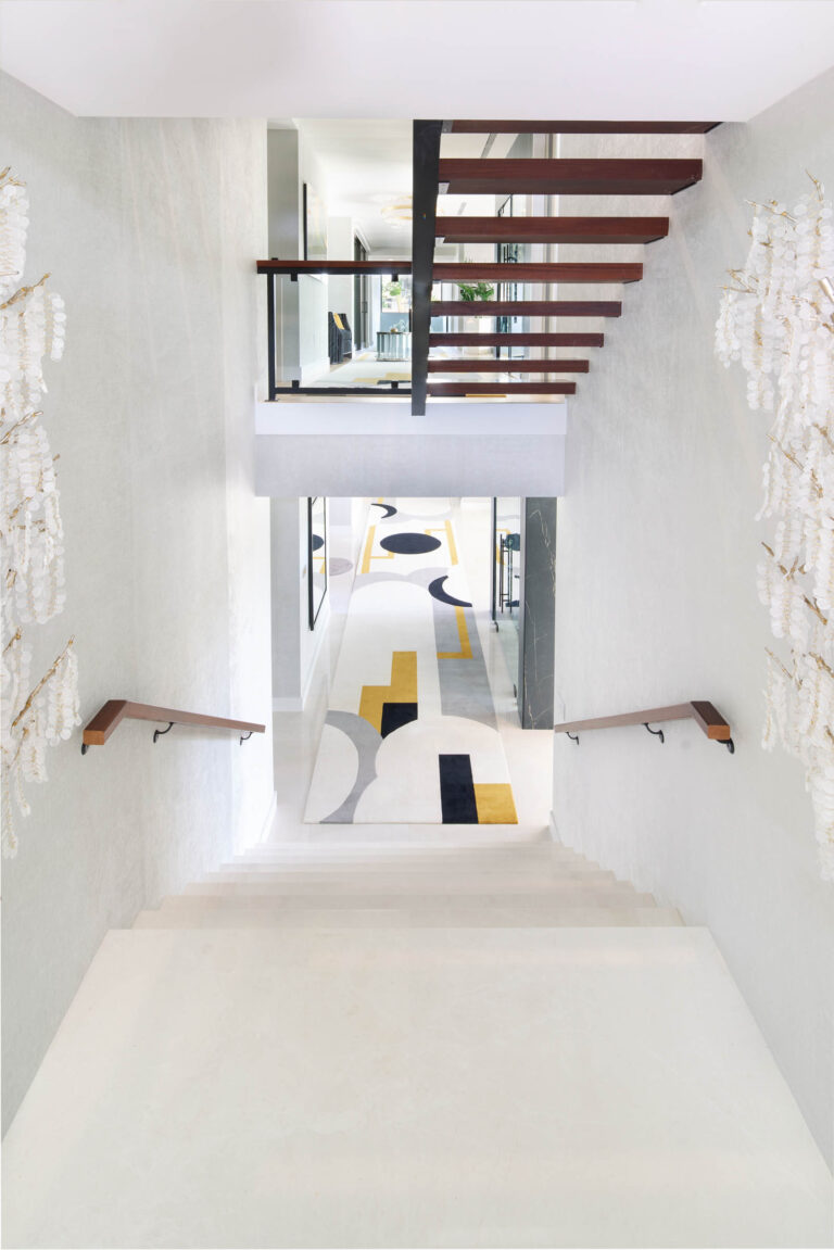 A modern staircase in a bright, spacious interior. The stairs feature wooden steps with sleek metal railings. Two walls are adorned with cascading artistic elements. At the bottom, a geometric-patterned carpet in white, yellow, and black colors leads into a hallway with framed artworks.