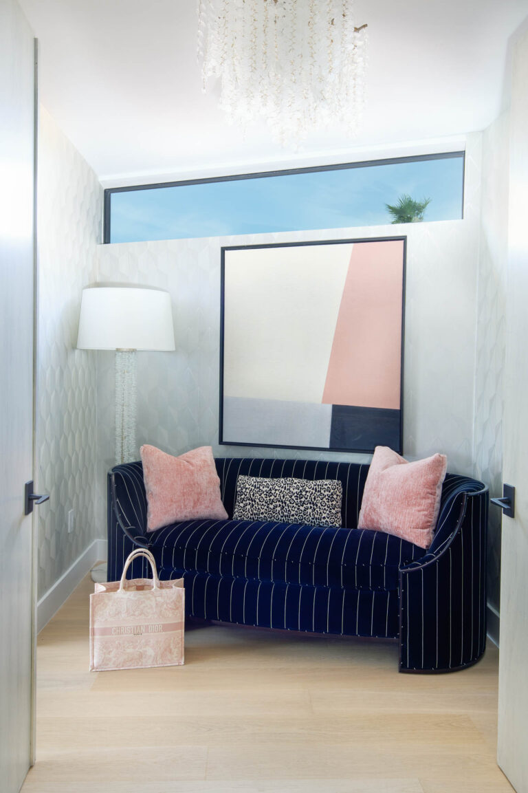 A cozy room featuring a navy striped sofa with pink and patterned cushions. A large abstract painting hangs above the sofa. Theres a tall floor lamp to the left and a delicate chandelier overhead. A pink tote bag sits on the floor. A window offers a glimpse of a clear blue sky.