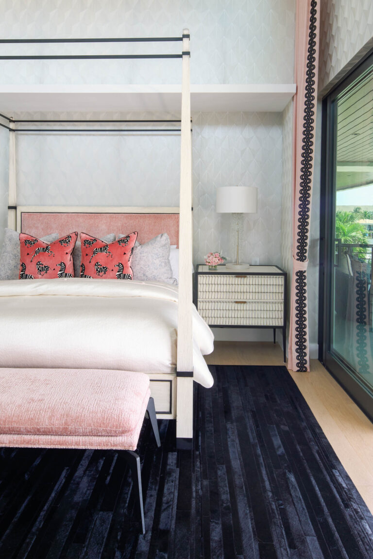 A stylish bedroom features a canopy bed with a pink cushioned bench at the foot. The bed has white and pink patterned pillows. A textured, light-colored wall runs behind a sleek, light wood nightstand with a white lamp. Floor-to-ceiling glass doors reveal a partial outdoor view. A dark rug lies on the wooden floor.